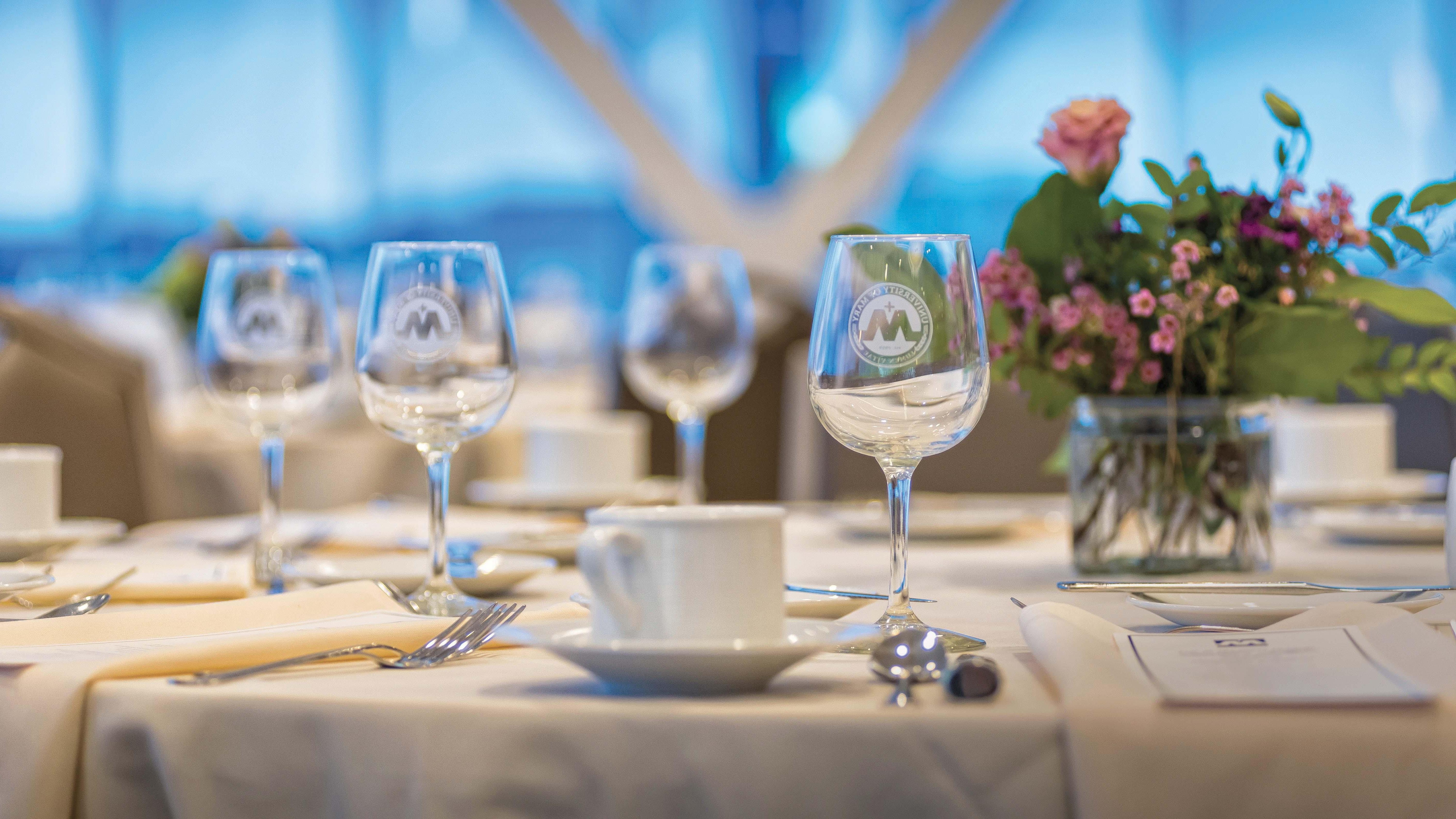 University of Mary wine glasses etched with its seal are set on a table with a centerpiece, 菜单, 银器, and napkins adding elegance to the formal dining decor inside 创始人霍尔 on campus.